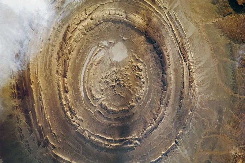 Concentric brown ridges of the Richat Structure seen from the International Space Station, with a pale patch of sand near the centre and cloud at upper left